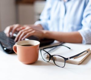 close-up view of person sitting at desk with computer, coffee and glasses