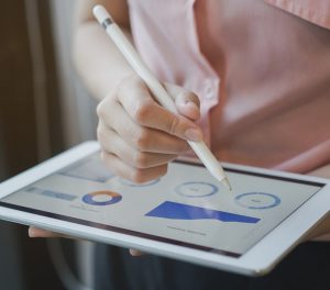 A woman's hand holding a stylus pointing to an ipad with charts and graphs