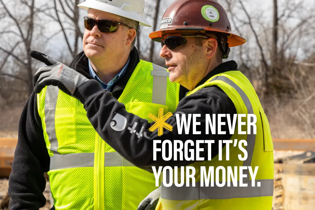 Two people in hard hats and safety vests are standing outside and discussing a project; the man on the right is pointing and both are looking to the left.