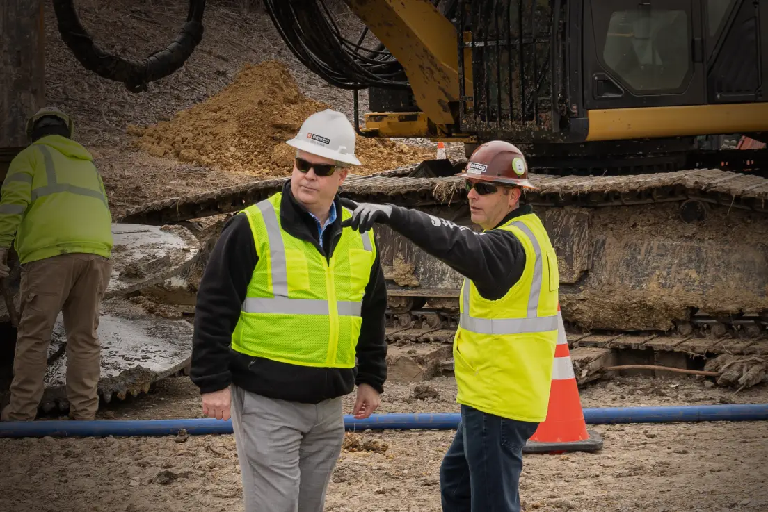 Two people in hard hats and safety vests are standing outside and discussing a project; the man on the right is pointing and both are looking to the left.