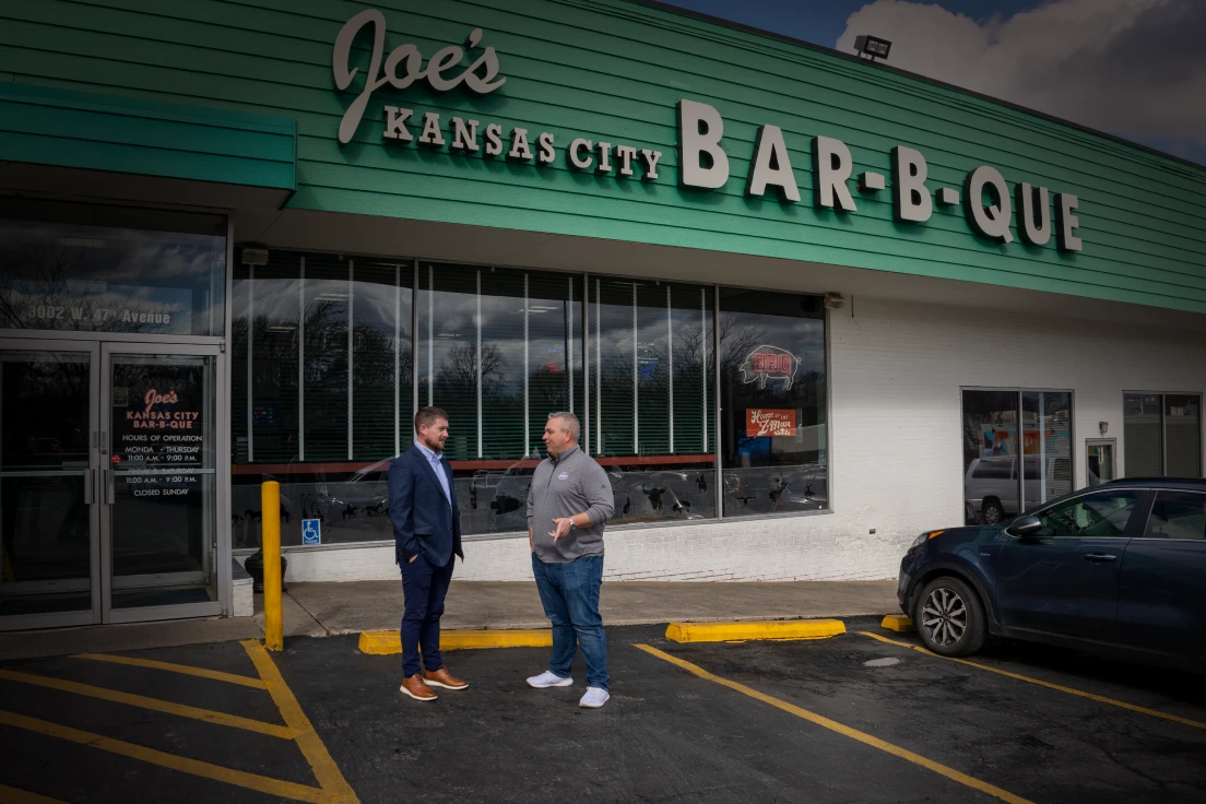 Two people stand and talk outside the front door of a Joe's Kansas City Bar-B-Q.