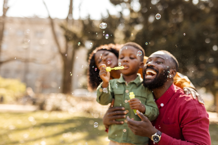 Father and young son blowing bubbles together in a sunlit park, both smiling.