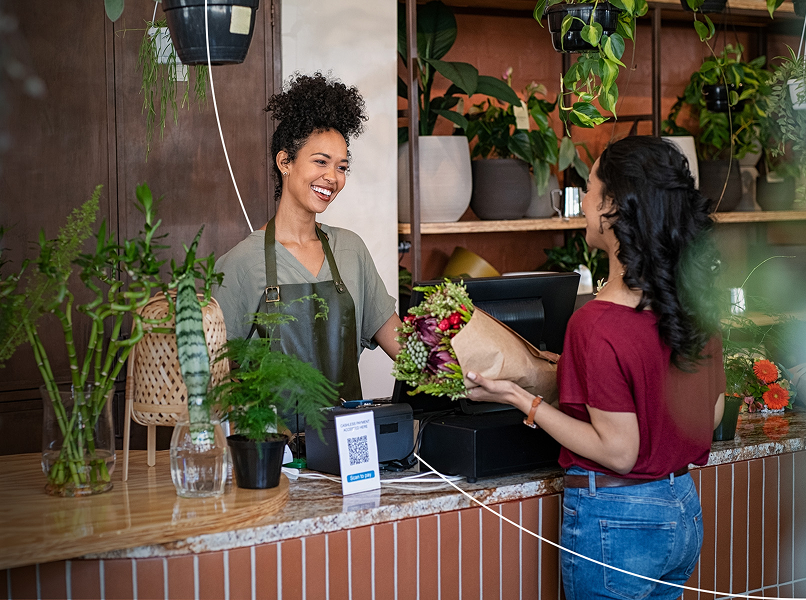 Smiling florist in an apron hands a bouquet to a customer at a wooden counter, surrounded by potted plants and greenery.