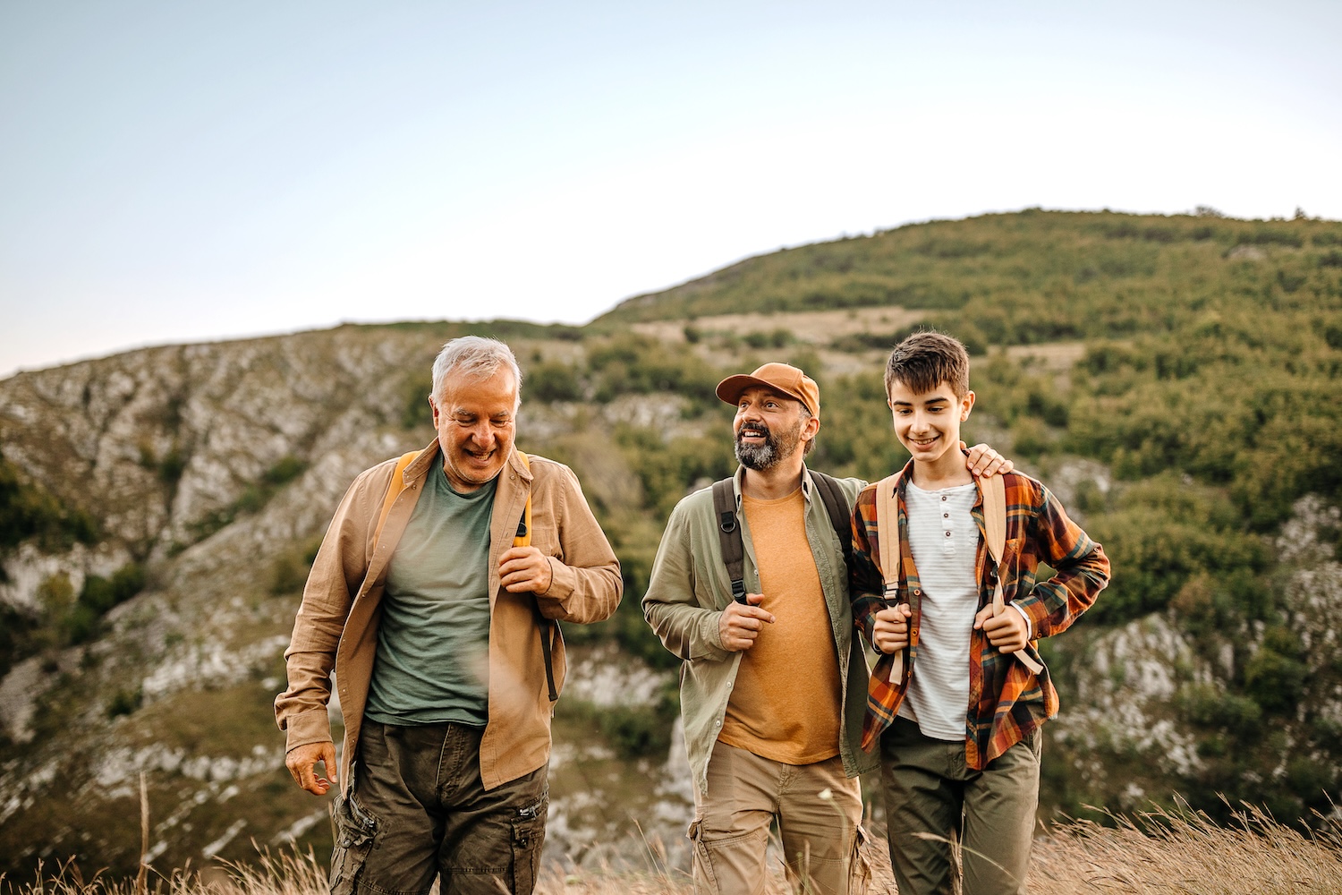 Three men of varying ages walking together outdoors with hills in the background.