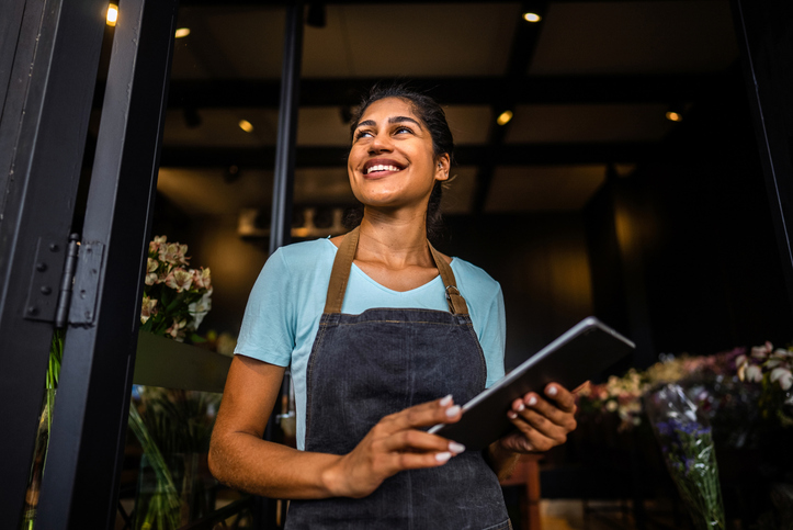 Shop attendant in a denim apron holding a tablet, smiling at the shop entrance with flowers in the background