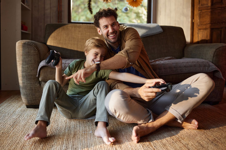 Father and son sit on the living room rug, playing a video game together with controllers and smiling at the screen.