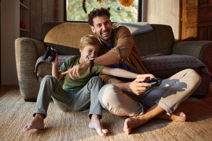 Father and son sit on the living room rug, playing a video game together with controllers and smiling at the screen.