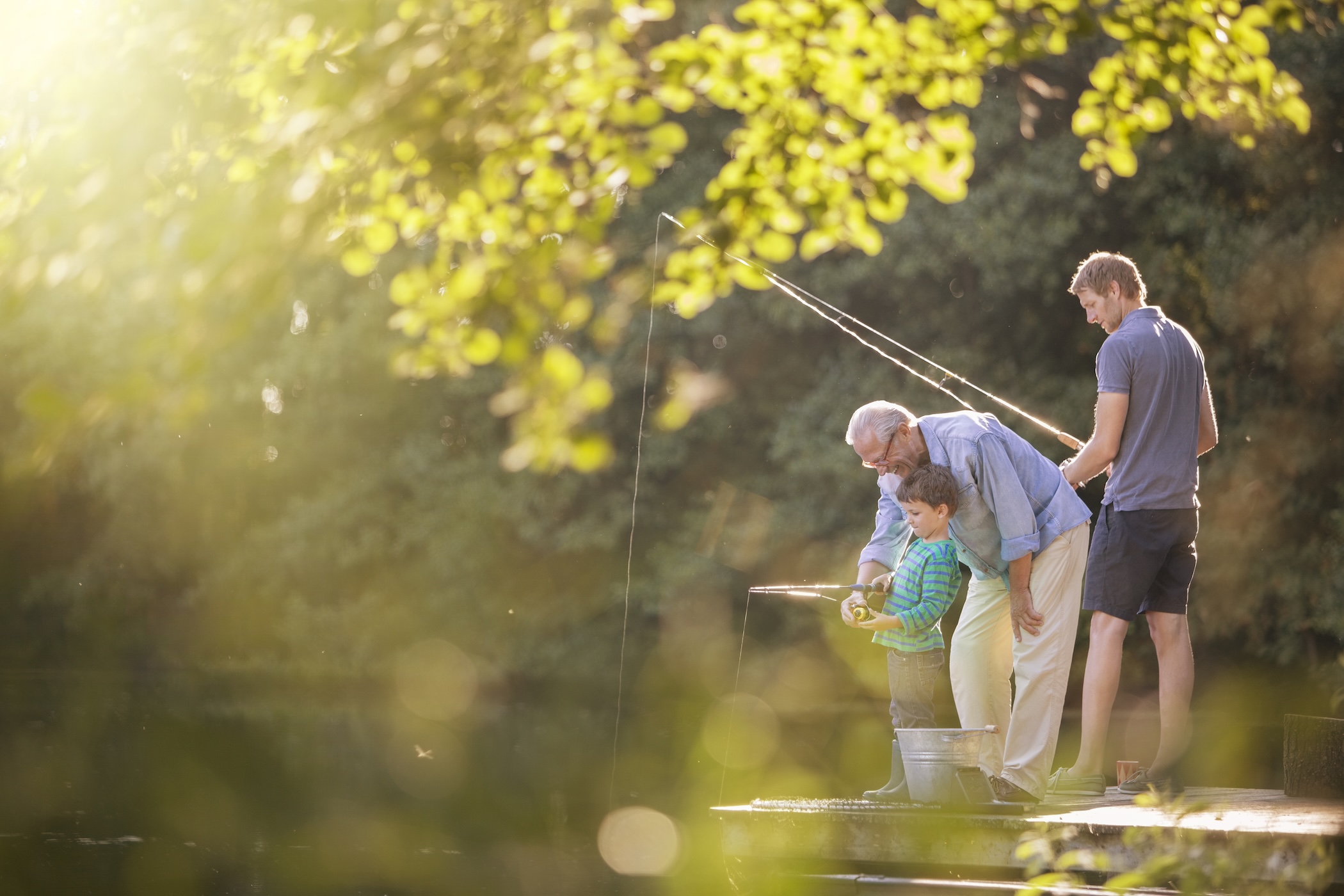 A grandfather, father, and young son fishing together near a sunlit pond.