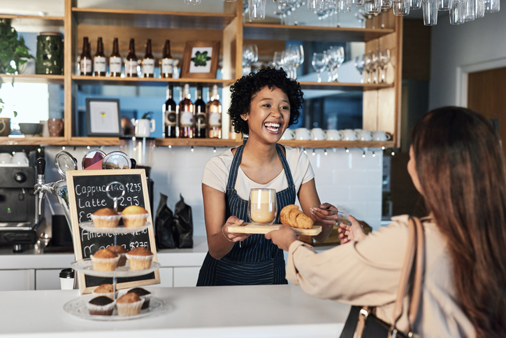 Smiling barista with curly hair hands a mug and pastries to a customer at a bright cafe counter, with muffins on a stand nearby.